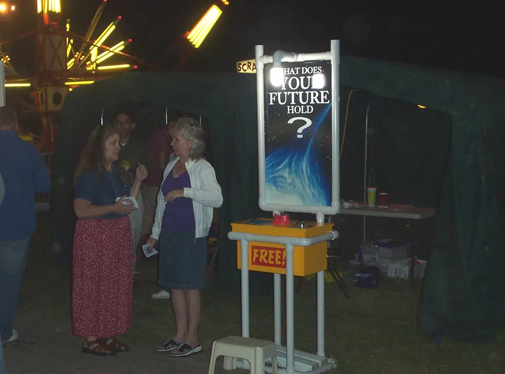 The fair booth at night under the lights of the rides, a conversation going on beside the sign