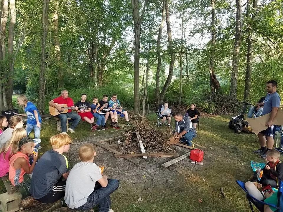 Campers seated in a circle around a campfire in the woods while a leader plays guitar